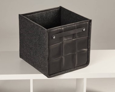 Keçe ve deriden yapılmış boş kare kutu, beyaz bir rafta dinleniyor, modern ve zarif bir depolama çözümü sunuyor.