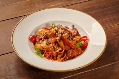 Rendelenmiş peynir ve otlarla süslenmiş et ve sebzelerle dolu pappardelle, ahşap bir masada beyaz bir tabakta servis ediliyor.