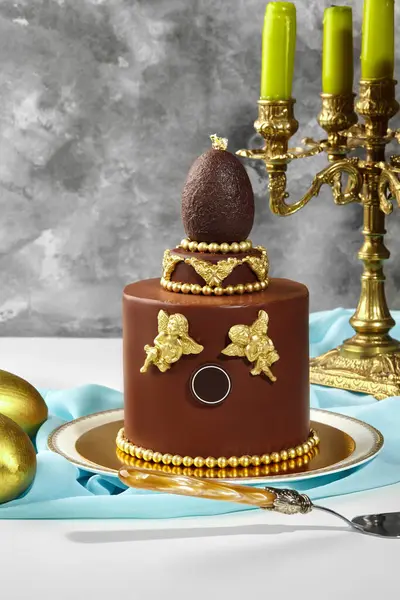 Altın melekler, çikolata yumurtası ve altın boncuklarla süslenmiş, altın bir mum tutacağının yanında duran lüks çikolatalı paskalya pastası.