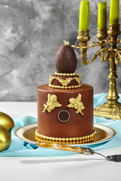 Altın melekler, çikolata yumurtası ve altın boncuklarla süslenmiş lüks çikolatalı paskalya pastası, yanında bir pasta sunucusu ve altın paskalya yumurtası olan zarif bir tabakta duruyor.