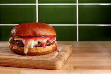 Erimiş peynirli ve ahududu soslu hamburger ahşap bir tahtaya damlıyor, yeşil fayanslı duvarın önünde.