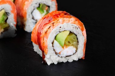 Avokado, krem peynir ve somonla dolu Uramaki suşi rulosu, üzerine sos serpiştirilmiş ve koyu bir arka planda sunulmuştur.