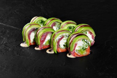 Somon, ton balığı, avokado, salatalık ve krem peynirli Uramaki suşisi, üzerine sos serpiştirilmiş siyah bir levha üzerine uzanır.