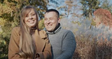 Gülümseyen ve kameraya bakan mutlu bir çiftin portresi. Sonbahar parkında kalın giysiler içinde dikiliyorum..