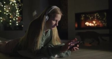 Kulaklıklı bir kız akıllı telefon kullanıyor. Şöminenin yandığı bir odada yerde yatıyor ve büyük bir Noel ağacı var. 4k video