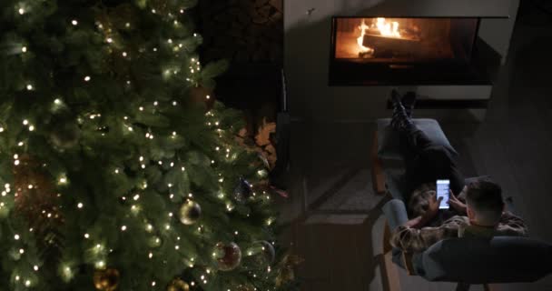 一个年轻人正在壁炉边和圣诞树旁休息 他用的是智能手机 从上面看 — 图库视频影像