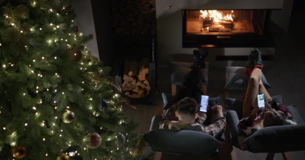 一对年轻夫妇正在壁炉边休息 还有一棵大圣诞树 使用智能手机在网上订购礼物 — 图库视频影像