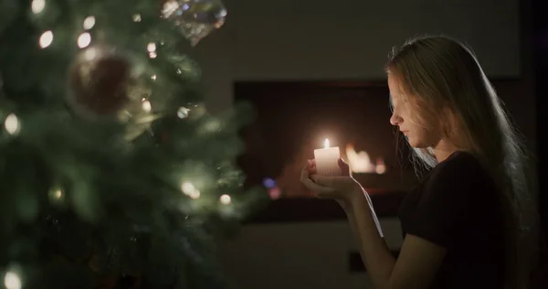 一个手里拿着蜡烛的女孩坐在圣诞树旁 在背景中 火在壁炉中燃烧 平安夜侧视图 — 图库照片