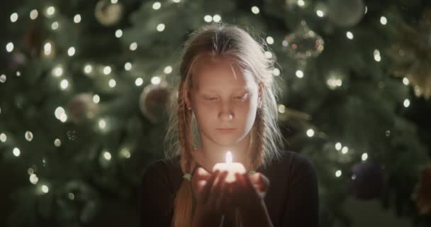 子供は手に火のろうそくを見ている ぼやけたクリスマスツリーのライトの背景に 願い事をする — ストック動画