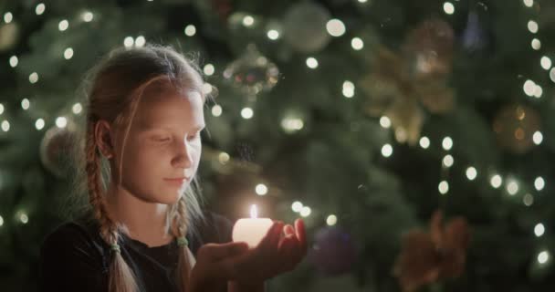 子供は手に火のろうそくを見ている ぼやけたクリスマスツリーのライトの背景に 願い事をする — ストック動画