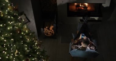 Bir adam şömine ve Noel ağacının yanında dinleniyor. Sıcak bir evde Noel arifesi.