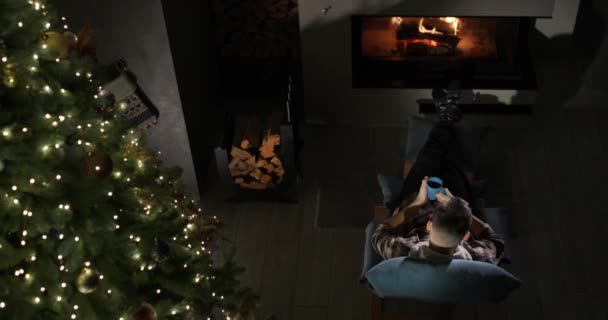 一个男人正在壁炉旁和圣诞树旁休息 平安夜在舒适的房子里 — 图库视频影像