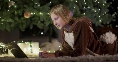 Geyik kostümlü bir genç evdeki büyük bir Noel ağacının arka planında dizüstü bilgisayar kullanıyor..