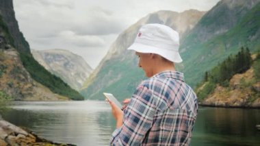 Bir turist bir smartphone pitoresk Norveç fiyordu arka plan üzerinde kullanır, uzaktan bir yolcu gemisi görünür. İskandinavya'ya yolculuk. Her zaman bağlantıda, teknoloji yolda. 4k video