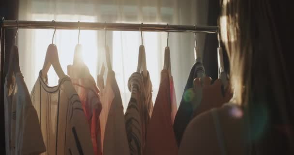 一个少女选择穿什么衣服 衣服挂在她的肩上在她面前 阳光照射着衣服 — 图库视频影像