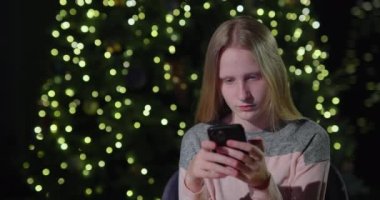 Genç bir kız Noel ağacının arka planında akıllı telefon kullanıyor..