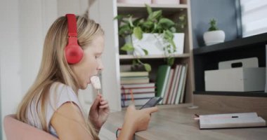 A teenager in headphones uses a smartphone, eats ice cream in his room.