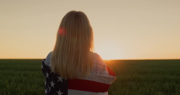 肩にアメリカ国旗の女性が太陽が小麦の畑に沈むのを見ている — ストック動画