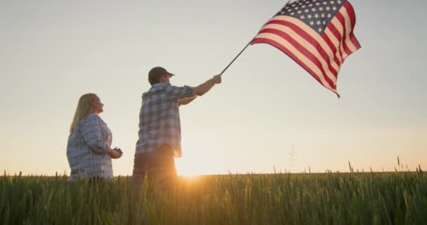 太陽が沈むと 小麦の畑の前でアメリカ国旗を振っている精力的なカップル 7月4日のコンセプト — ストック動画