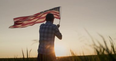 Amerikan bayrağını sallayan genç bir adamın silueti. Gün batımında bir buğday tarlasında durmak.