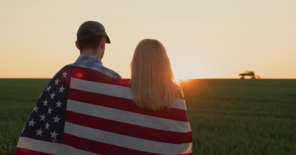 トラクターが遠くで働いている小麦畑を見ています彼らの肩にアメリカの旗を持つ農家のカップルは — ストック動画