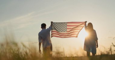Bir adam ve bir kadın güneş batarken buğday tarlasının üzerinde Amerikan bayrağını göndere çekiyor. ABD 'nin bağımsızlık günü kavramı.
