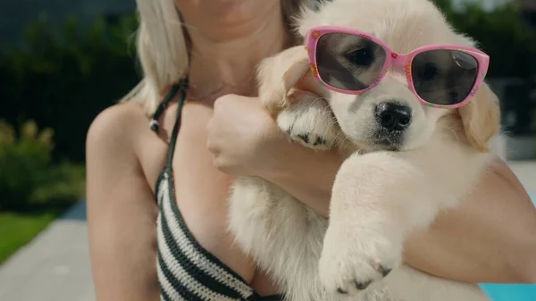 A woman in a bikini holds a blonde puppy in sunglasses in her arms. Summer and vacation with a pet concept.