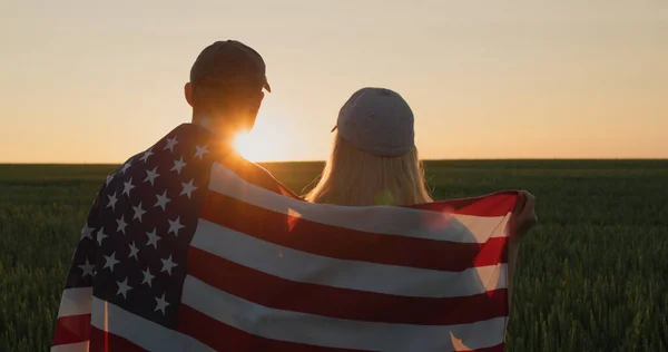 A man and a woman with the US flag on their shoulders look at the sunrise over a field of wheat.