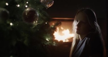 Bir çocuk karanlıkta bir Noel ağacının yanında oturuyor, arka planda bir şöminede ateş yanıyor. Noel Arifesi.