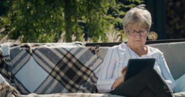 Tablet kullanan yaşlı bir kadının portresi. Bir evin arka bahçesinde salıncakta sallanıyor..