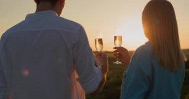 Kırmızı şarap kadehleriyle genç çift resimli bir vadide gün batımını seyrediyor..