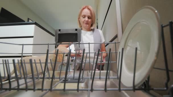 一位中年妇女把盘子放在洗碗机里 旁边放着她的狗 宠物陪伴下的家务活 — 图库视频影像