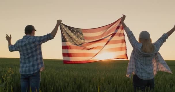 幸せなカップルは 日没時に小麦畑の背景に立って 米国の国旗を上げます — ストック動画