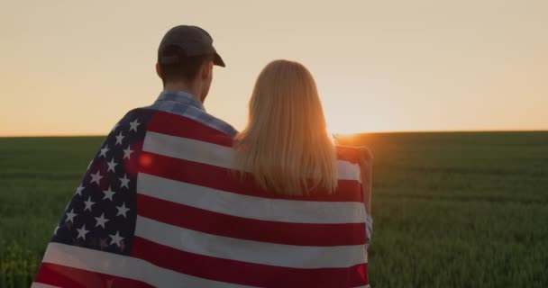小麦畑の日の出を賞賛する肩に米国旗を持つ若いカップル — ストック動画