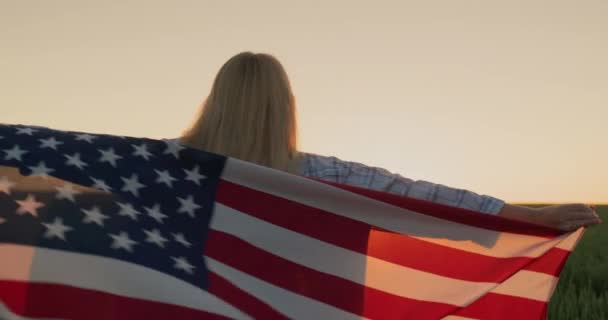 一个肩上扛着美国国旗的女人看着太阳落在麦田上 — 图库视频影像