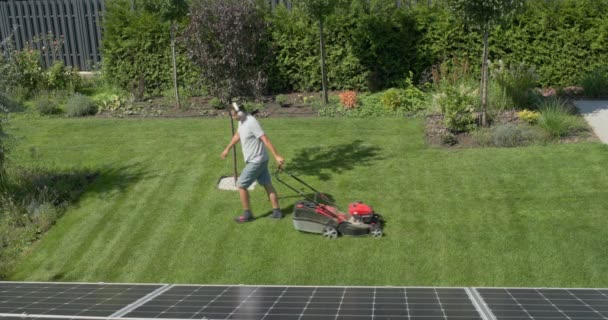 中年の男が家の裏庭で芝生を刈る ヘッドフォンで音楽を聴く — ストック動画