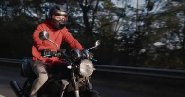 Kırmızı ceketli bir adam ormanda motosiklet sürüyor, güneş yüzünü aydınlatıyor..