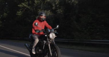 Kırmızı ceketli bir adam ormanda motosiklet sürüyor, güneş yüzünü aydınlatıyor..
