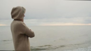 A woman in a warm knitted sweater looks at the sea where the storm begins.