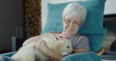 Yaşlı bir kadın kucağında bir köpek yavrusuyla bir sandalyede dinleniyor. Ev konforu ve güvenli yaşlılık.