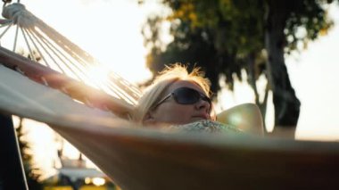 Yaz elbise giymiş bir kadın turist bir hamakta rahatlatır. Ufuk güneş güzel parlak nokta çerçevede oluşturur. 4k video