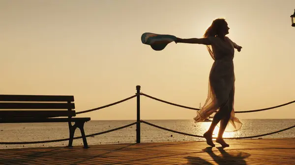 女性は 海で夜明けを満たしています 感情的に桟橋に彼の手で帽子を回転しています 旅行の概念の夢 ストック画像