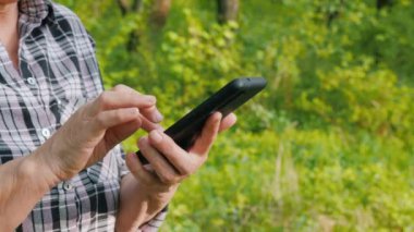 Parkta veya ormanda yürürken akıllı telefon kullanan yaşlı bir kadının elleri.