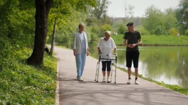 Yaşlı bir kadın parkta bir yürüyüşçünün yardımıyla yürüyor, kızı ve torunu da ona eşlik ediyor..
