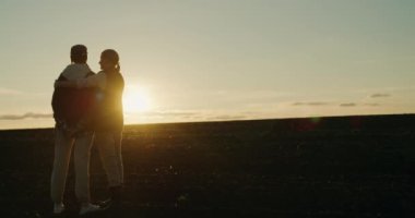 Sahada gün batımını seyreden bir kadın, genç kızını sevgiyle kucaklıyor..