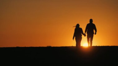 Genç bir çift, el ele tutuşarak batan güneşe doğru bir tarlada yürüyor..