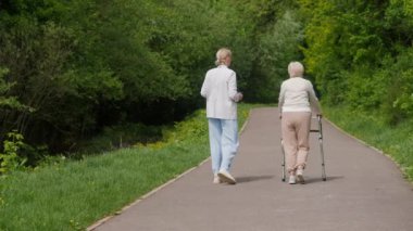 Yaşlı bir kadın bir yürüyüşçünün yardımıyla yürüyor, bir sağlık görevlisi de ona eşlik ediyor..
