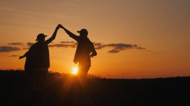 İki çiftçi bir tarlada neşeyle dans ediyor. Siluetler güneşin battığı gökyüzüne karşı.