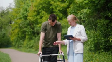 Orta yaşlı bir kadın, yemyeşil bir parkta tabletle meşgul olurken, yürüteç kullanan genç bir adama yardım ediyor. Bu görüntü teknolojinin rehabilitasyondaki rolünü ve desteğin önemini vurgular.