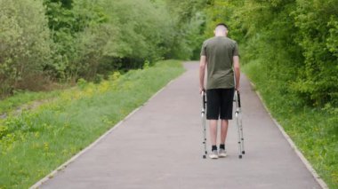 Genç bir adam yemyeşil bir parkta yol boyunca yürüyen birini kullanıyor. Sahne, hareketlilik yardımlarının, esnekliğin ve açık hava faaliyetlerinin iyileşme ve iyileşme için yararlarının önemini vurguluyor.
